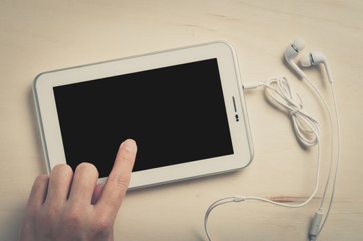 Young man left hand touching on small tablet pc screen on wood table with in ear headset beside with vintage filter effect