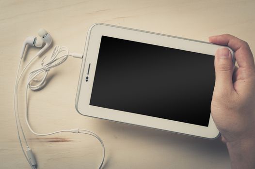 Young man hand holding small tablet pc screen on wood table with in ear headset beside with vintage filter effect