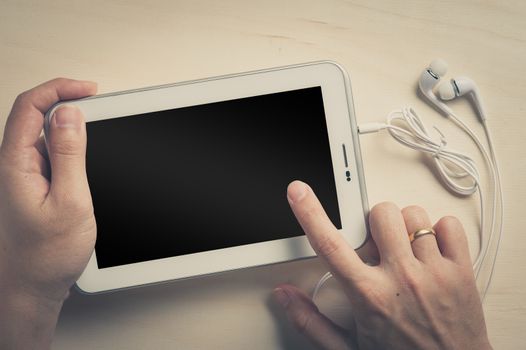 Young man hand touching on small tablet pc screen on wood table with in ear headset beside with vintage filter effect