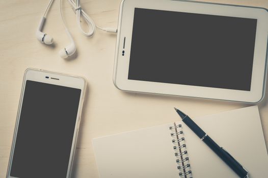 Small tablet pc with blank area on touchscreen, smart phone, in ear headset, notebook, and pen on wood table with vintage filter effect