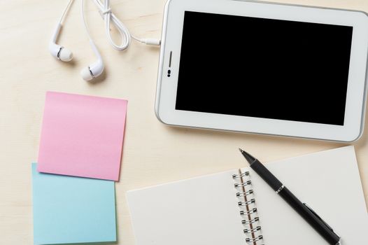 Small tablet pc with blank area on touchscreen, in ear headset, notebook, sticky paper, and pen on wood table