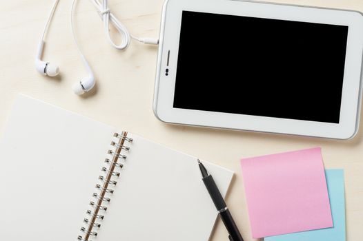 Small tablet pc with blank area on touchscreen, in ear headset, notebook, sticky paper, and pen on wood table
