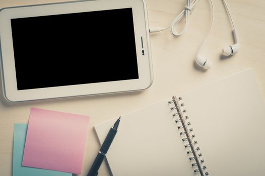 Small tablet pc with blank area on touchscreen, in ear headset, notebook, sticky paper, and pen on wood table with vintage filter effect
