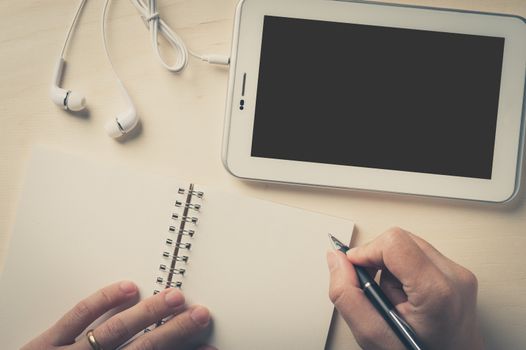 Young man hand writing on notebook beside small tablet pc and in ear headset on wood table with vintage filter effect