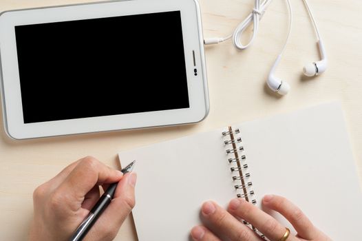 Young man hand writing on notebook beside small tablet pc and in ear headset on wood table