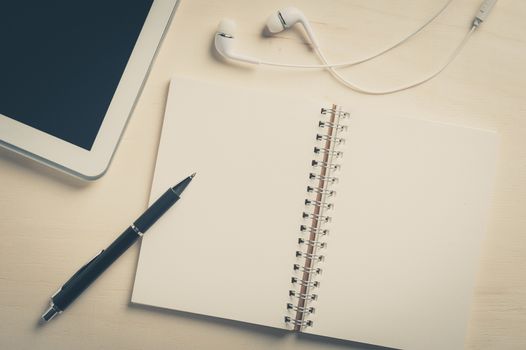 Notebook with blank area for text or message and pen beside small tablet pc and in ear headset on wood table with vintage filter effect