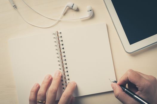 Young man right hand writing on notebook beside small tablet pc and in ear headset on wood table with vintage filter effect