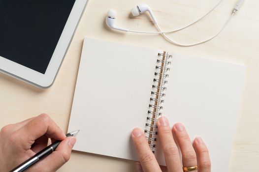 Young man left hand writing on notebook beside small tablet pc and in ear headset on wood table