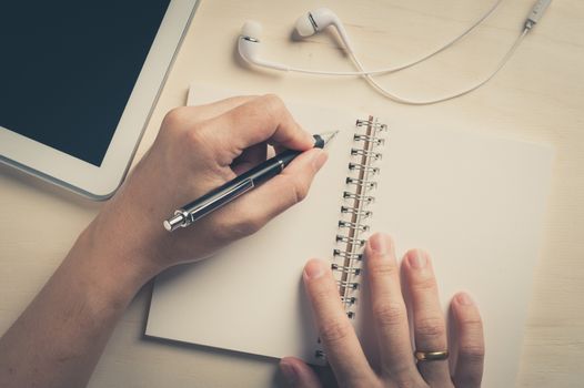 Young man left hand writing on notebook beside small tablet pc and in ear headset on wood table with vintage filter effect