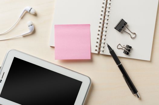 Sticky paper with blank area for text or message, opened notebook,and pen beside small tablet pc and in ear headset on wood table