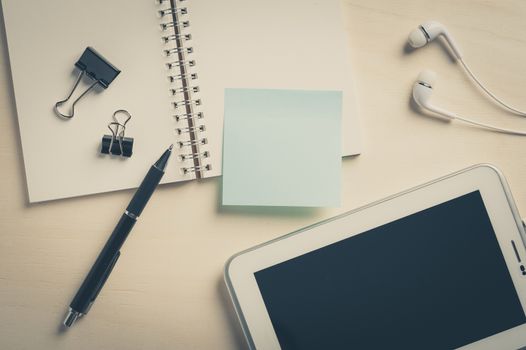 Sticky paper with blank area for text or message, opened notebook,and pen beside small tablet pc and in ear headset on wood table with vintage filter effect