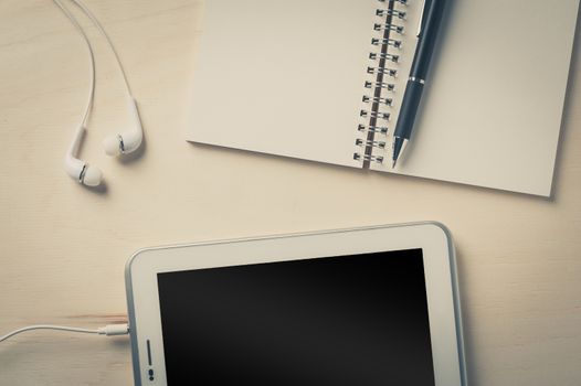 Notebook, pen, small tablet pc, and in ear headset on wood table with vintage filter effect