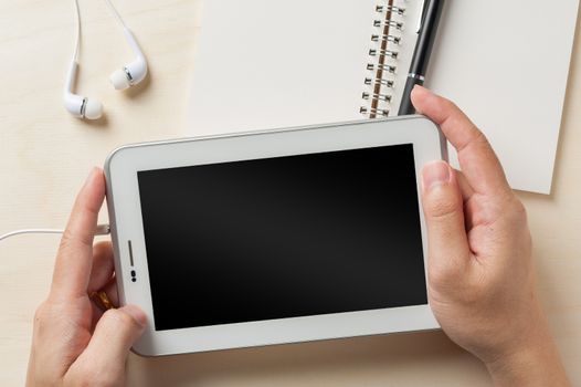 Young man hands holding small tablet pc screen on wood table with in ear headset and notebook beside