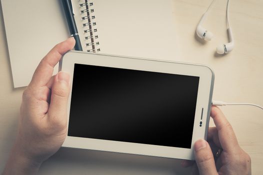 Young man hands holding small tablet pc screen on wood table with in ear headset and notebook beside with vintage filter effect