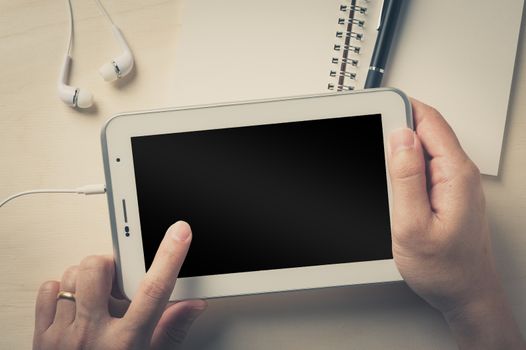 Young man left hand touching on small tablet pc screen on wood table with in ear headset and notebook beside with vintage filter effect