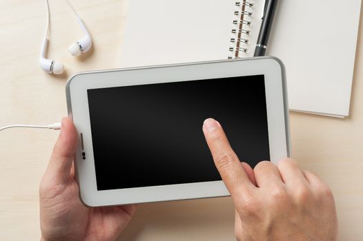 Young man right hand touching on small tablet pc screen on wood table with in ear headset and notebook beside