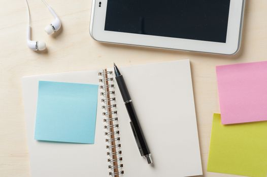Sticky paper with blank area for text or message, opened notebook,and pen beside small tablet pc and in ear headset on wood table