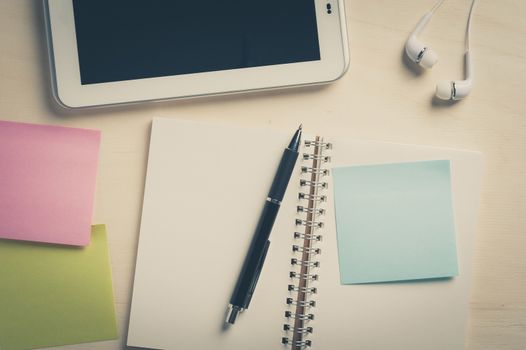 Sticky paper with blank area for text or message, opened notebook,and pen beside small tablet pc and in ear headset on wood table with vintage filter effect