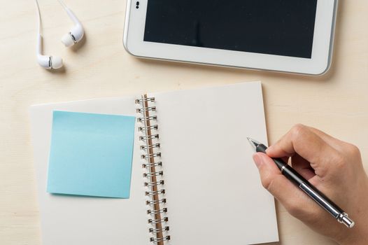 Young man right hand writing on notebook with sticky paper beside small tablet pc and in ear headset on wood table