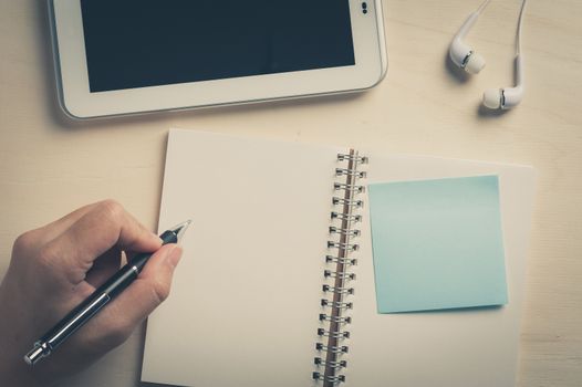 Young man left hand writing on notebook with sticky paper beside small tablet pc and in ear headset on wood table with vintage filter effect