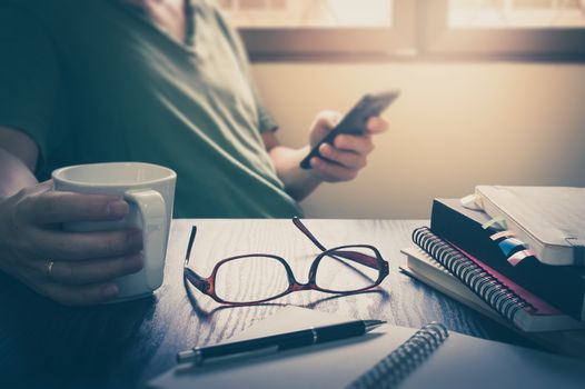 Glasses put down on table beside notebooks and pen while young freelance worker using smart phone and holding coffee cup in morning time on work day, Working at home concept with vintage filter effect