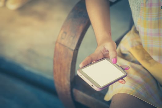 Young hipster woman holding smartphone on her right hand while sitting on wood bench in morning time with vintage filter effect