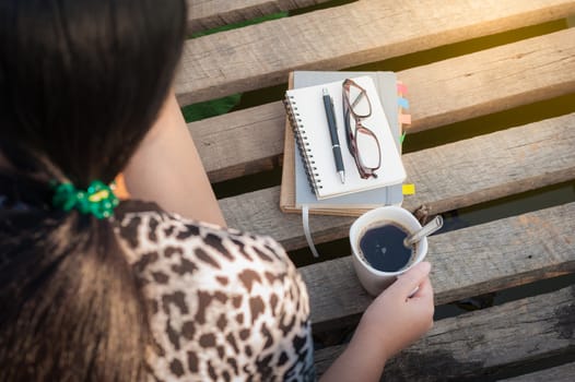 Young woman holding coffee cup while sitting on wood bridge with notebooks, pen, and glasses in morning time on weekend