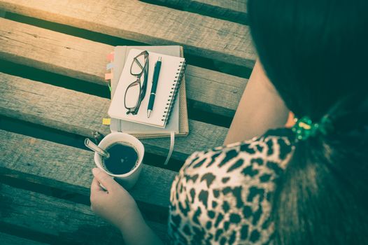 Young hipster woman holding coffee cup while sitting on wood bridge with notebooks, pen, and glasses in morning time on weekend with vintage filter effect