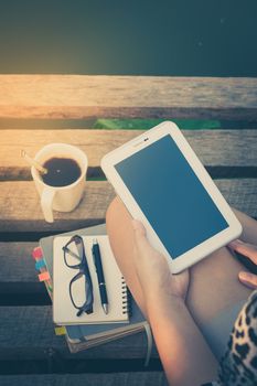 Young woman holding small talet with left hand while sitting on wood bridge with coffee cup, glasses, and notebooks in morning time on weekend with vintage filter effect