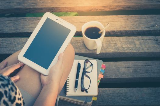 Young woman holding small talet while sitting on wood bridge with coffee cup, glasses, and notebooks in morning time on weekend with vintage filter effect