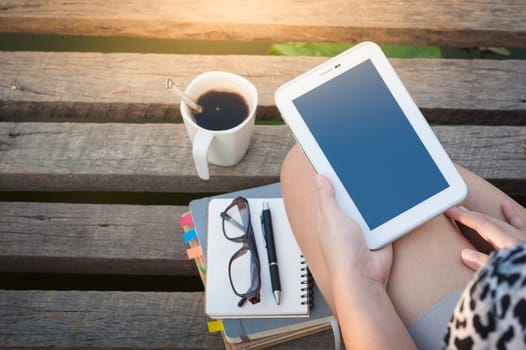 Young woman holding small talet with while sitting on wood bridge with coffee cup, glasses, and notebooks in morning time on weekend