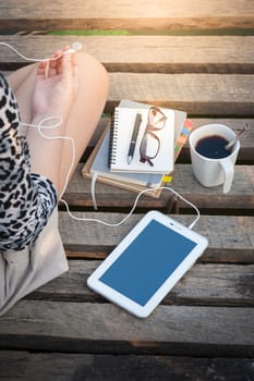 Young woman sitting on wood bridge with small tablet pc, coffee cup, glasses, and notebooks while listening to music in morning time