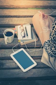 Young woman sitting on wood bridge with small tablet pc, coffee cup, glasses, and notebooks while listening to music in morning time with vintage filter effect