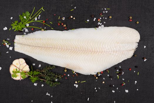 Fresh halibut fillet with fresh herbs and garlic on dark black stone background top view. Luxurious seafood eating background. 