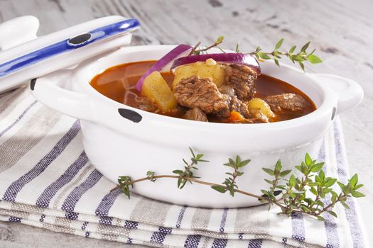 Delicious goulash soup in white pot on white wooden background. Culinary traditional goulash eating.