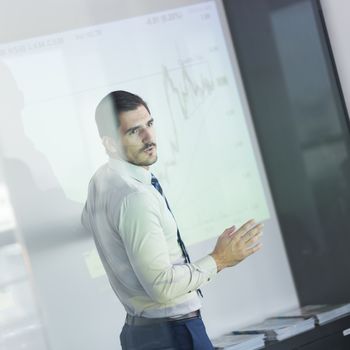 Business man making a presentation in front of whiteboard. Business executive delivering a presentation to his colleagues during meeting or in-house business training. View through glass.