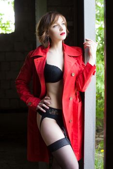 Attractive young woman alluring in sexual lingerie and red coat at grunge industrial setting. Beauty, fashion. Concept: seduction, exhibitionism.