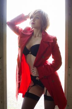 Attractive young woman alluring in sexual lingerie and red coat at grunge industrial setting. Beauty, fashion. Concept: seduction, exhibitionism.