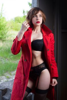 Attractive young woman alluring in sexual lingerie and red coat at grunge industrial setting. Beauty, fashion. Concept: seduction, exhibitionism.