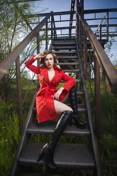 Attractive young woman alluring in sexual lingerie and red coat at grunge industrial setting. Beauty, fashion. Concept: seduction, exhibitionism.