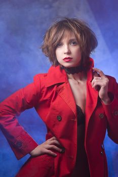 Attractive young woman alluring in sexual lingerie and red coat at smoke background. Beauty, fashion. Concept: seduction, exhibitionism.