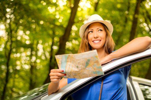 Young beautiful woman standing leaning at car in the forest, holding map and looking away.