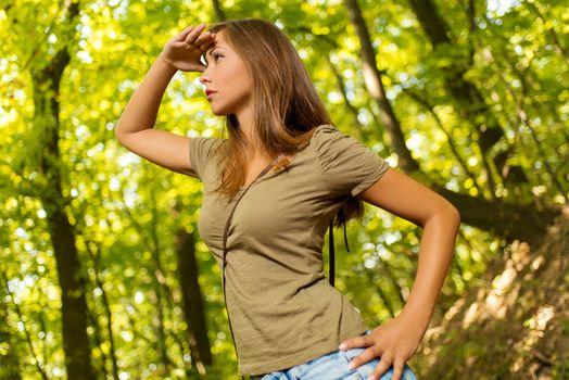 Beautiful young woman walking through forest and looking far away and covering sunlight his hand.