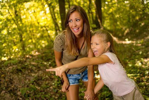 Beautiful young mother and her daughter walking through forest. Little girl pointing with finger far away.