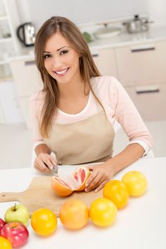 Cute young woman cutting grapefruit in kitchen. Looking at camera.