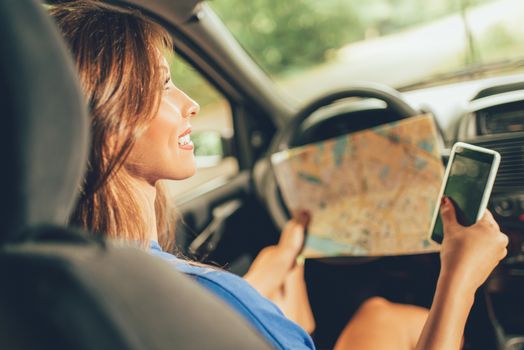 Young beautiful woman sitting in car holding map and using at smart phone. Rear view.
