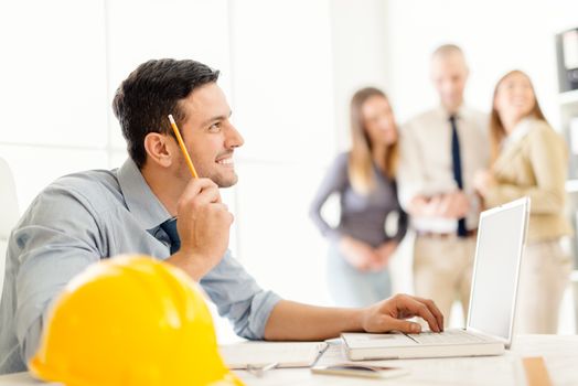 Young arabian male contractor sitting at laptop in office and thinking. His colleagues standing in background. 