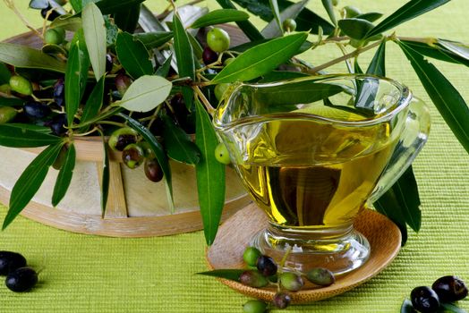 Olive Oil in Glass Gravy and Raw Green and Black Olives with Leafs in Wooden Bowl closeup on Green Textile background