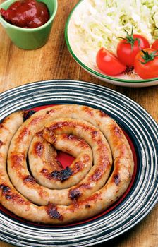 Arrangement of Delicious Grilled Spiral Sausage on Striped Plate with Ketchup, Cherry Tomatoes and Cabbage Salad closeup on Wooden Cutting Board