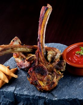 Arrangement of Roasted Lamb Ribs with Bread Sticks and Tomato Sauce on Stone Plate closeup on Dark Wooden background